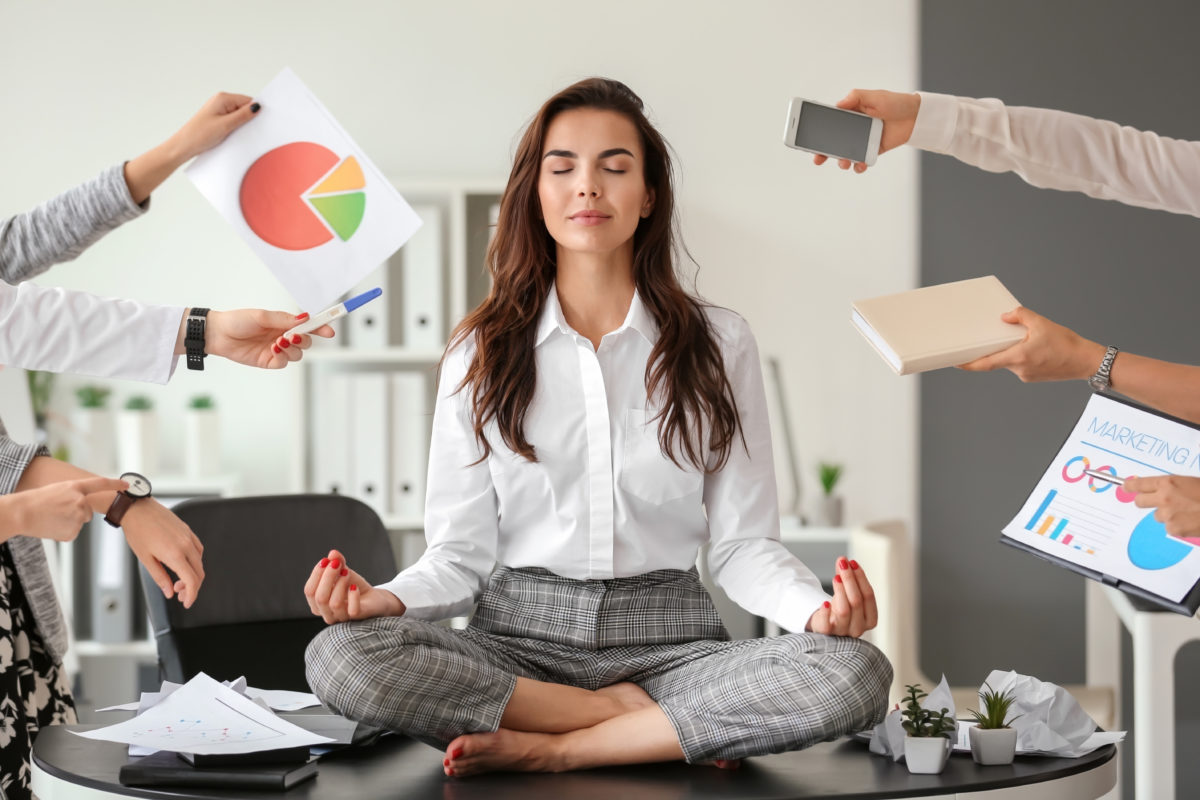 woman sitting on a desk meditating with people holding charts and work around her