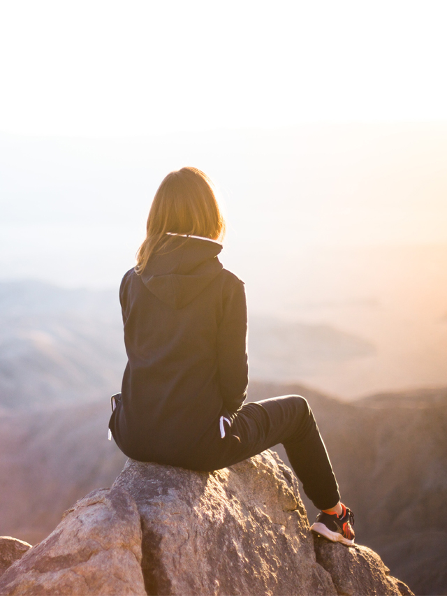 Woman sitting on a rock looking over the mountainous landscape as the poster image for the How to Balance Your Hormones Naturally web story