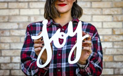 Woman in a flannel shirt holding up a cut out of the word "joy"